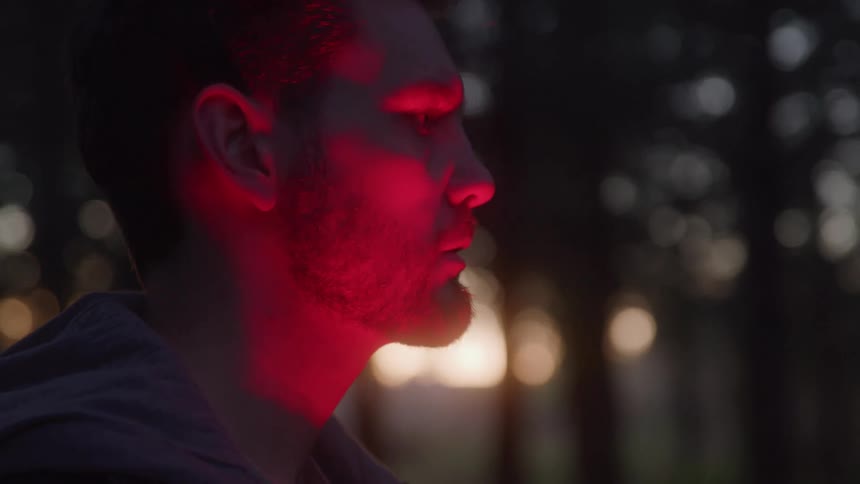 Man lit by red light stares intently at something in the forest at dusk