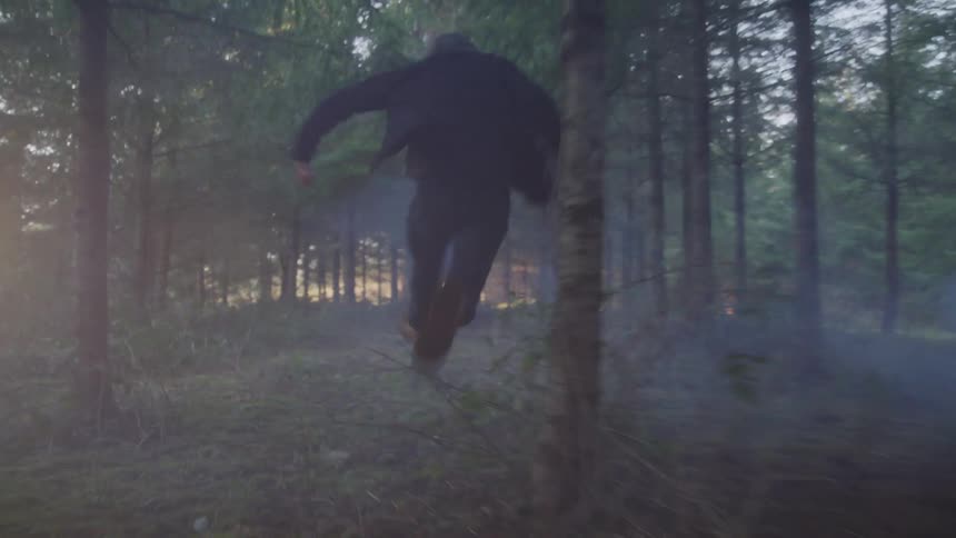A man wearing a jacket runs through the woods in a foggy atmosphere at midday