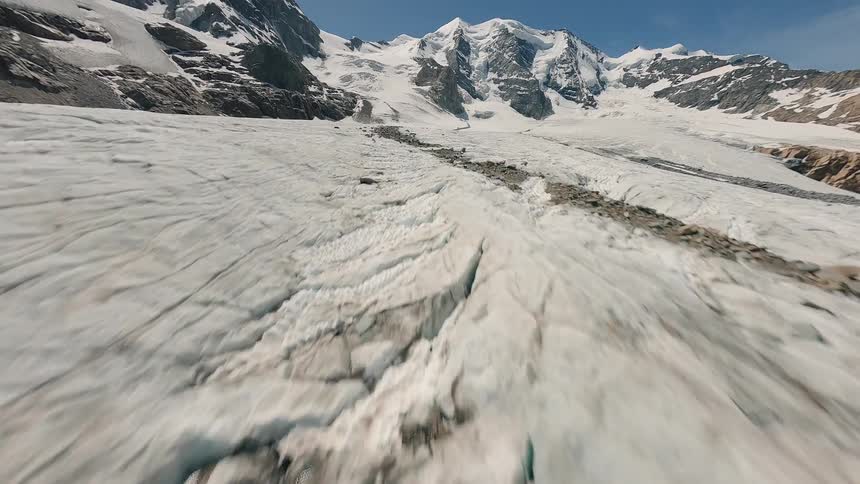 The video presents an aerial view of a vast rugged glacier with deep crevices and undulating ice formations as the camera pans upwards towering snowcapped mountains come into view under a clear sky revealing a majestic alpine landscape