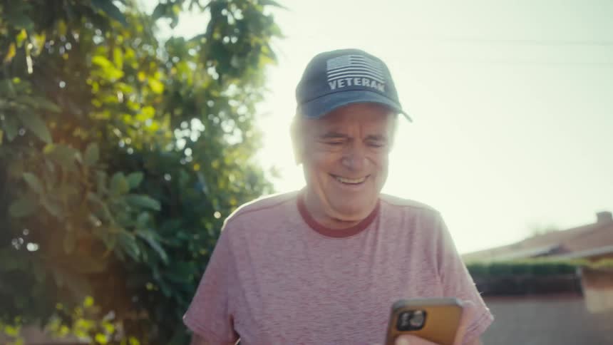The video features a smiling elderly man wearing a blue veteran baseball cap and a light pink tshirt he is holding a smartphone and looking down at it with bright sunlight shining from behind and through green foliage