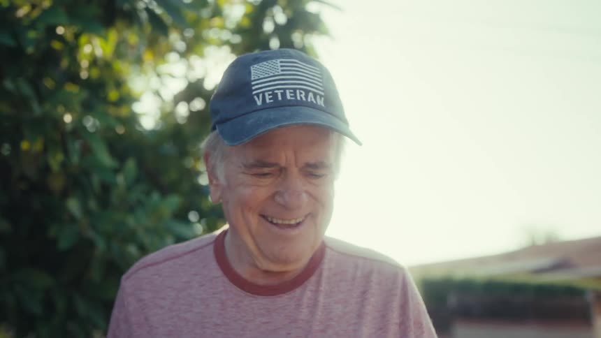An older man wearing a veteran cap smiles gently while walking in a tree lined neighborhood