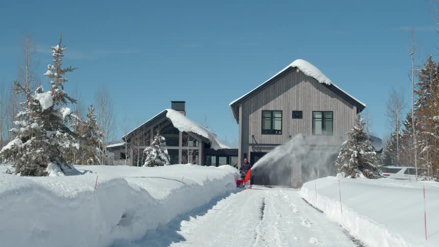 Man snowblowing a long driveway in front of a modern house