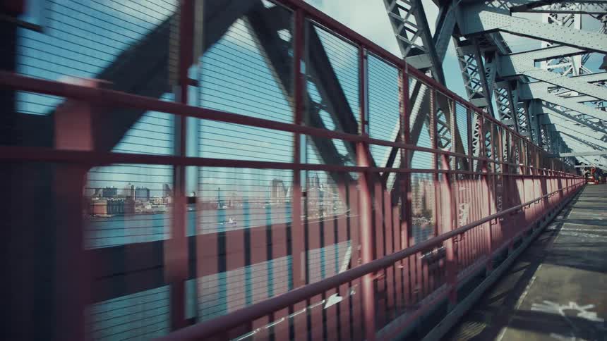A fenced pedestrian path runs alongside a steel bridge overlooking a calm river and city skyline in the distance