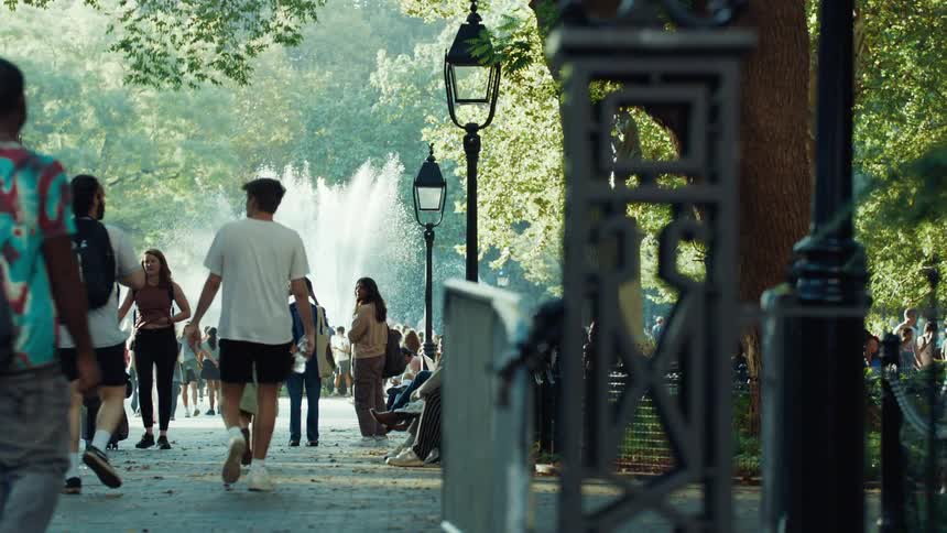 A busy park path is filled with numerous adults and young adults walking and sitting on benches among tall trees and old street lamps with a water fountain spraying in the background