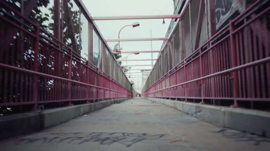 A wide pedestrian walkway with red metal fences on both sides extends into the distance with graffiti on the concrete ground and a view of buildings and trees above under an overcast sky
