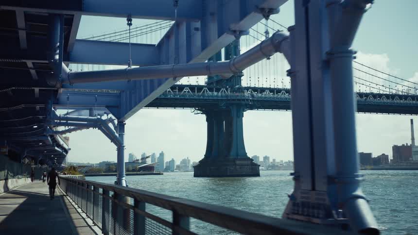 A young adult male walks along a pedestrian path under the structural beams of a large bridge with a wide river and a distant city skyline under a clear sky