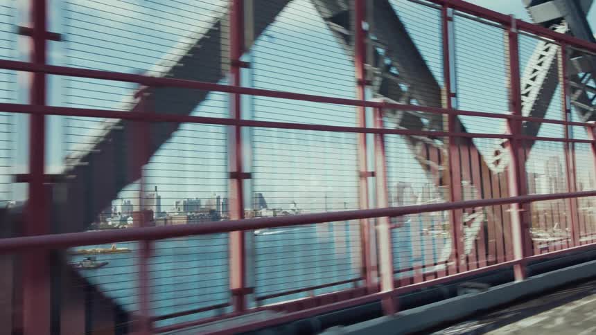 The video shows a view from what appears to be a moving vehicle or train looking out through the red girders of a bridge beyond the bridges structure a cityscape with numerous buildings stretches across the horizon with a body of water in the middle