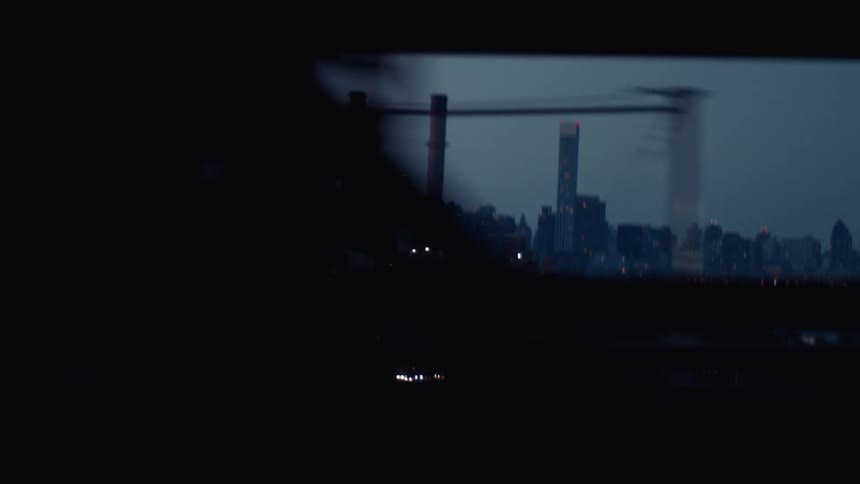 A dimly lit city skyline is seen through a moving frame with smokestacks and skyscrapers silhouetted against the twilight