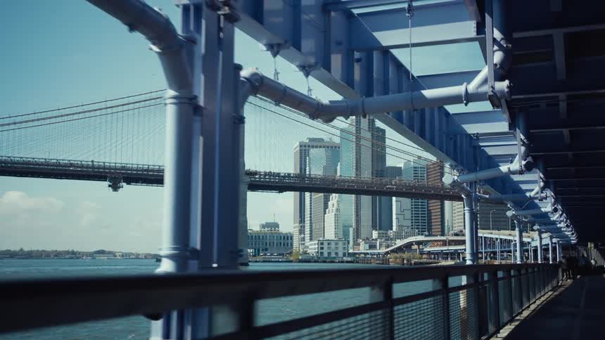 A bridge and buildings in the background all viewed from beneath the structural elements of a parallel elevated walkway on a clear day