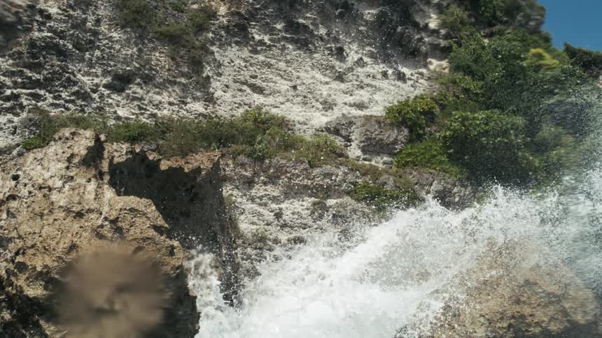 Ocean waves crash against a rocky island at midday causing splashes