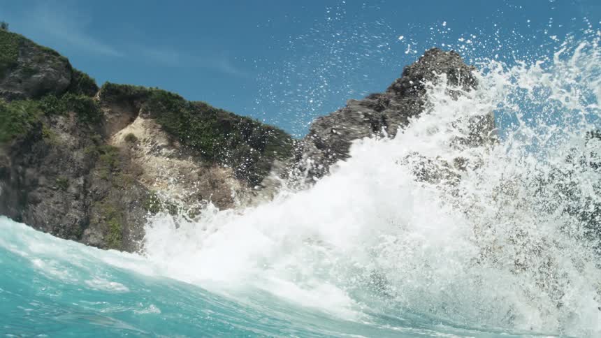 Turquoise ocean water crashes against a large textured rock formation sending white spray and droplets into the air with steep cliffs covered in green foliage and a clear blue sky in the background