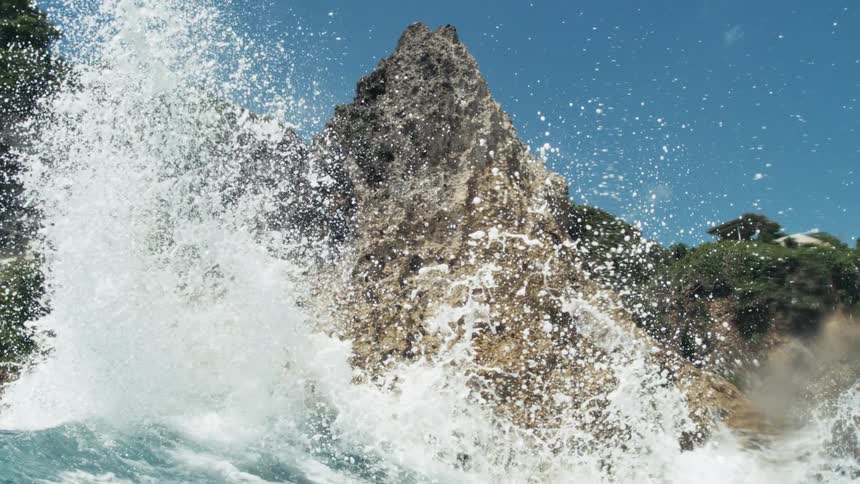 Waves crashing against a cliff side coastal rock formation sprays water into the air on a summer day