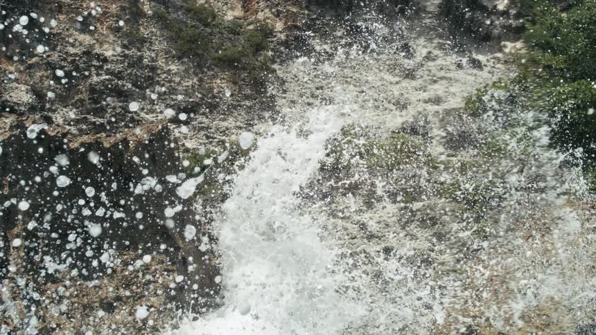 Ocean water violently crashes and sprays against rugged rock formations and cliffs with green foliage creating a dynamic scene of white foam and countless droplets flying through the air