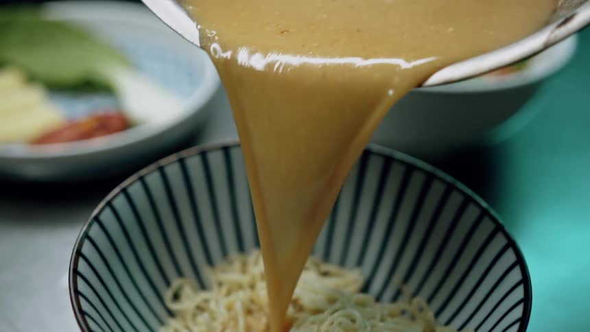 Thick orange brown sauce is poured from a metal pot into a striped bowl filled with noodles preparing a meal