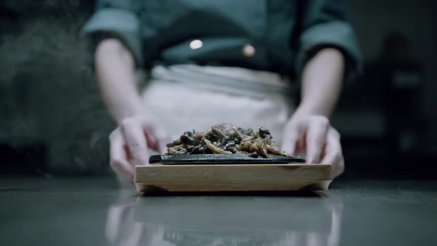 A chef presents a steaming hot plate of food on a wooden board with the dish placed at the center of a reflective counter
