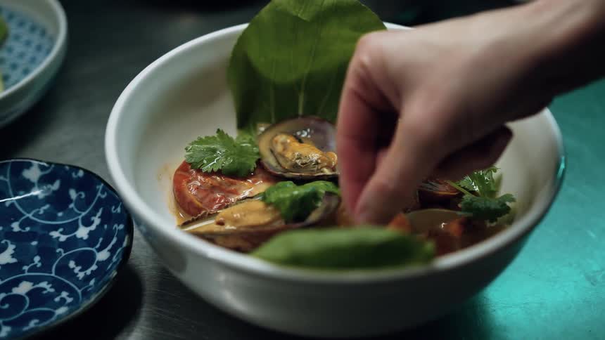A chefs hand meticulously garnishes a white bowl of mussels and tomatoes with fresh cilantro positioned on a metallic surface with other bowls in the blurred background