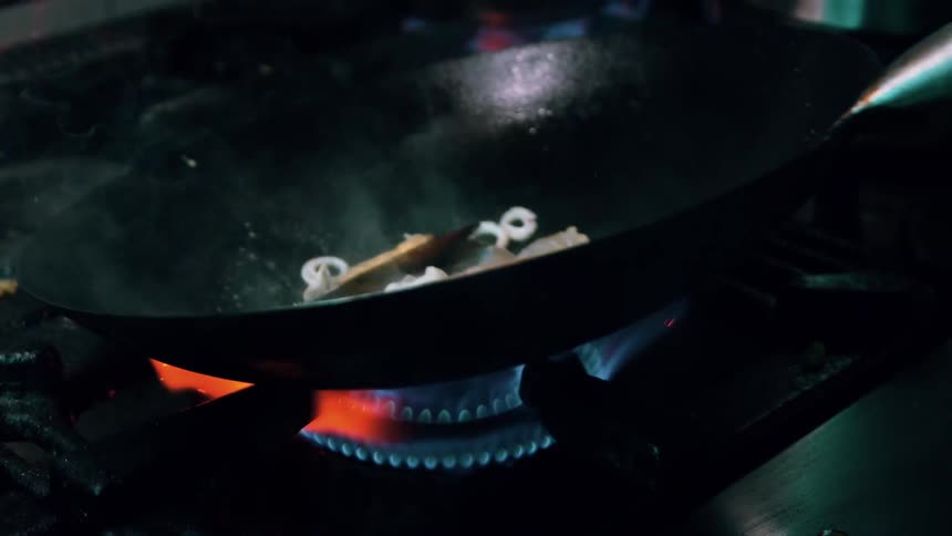 The video features a closeup of a wok on a gas burner where seafood including shrimp and calamari rings are being stirfried flames from the burner flare up around the wok indicating highheat cooking