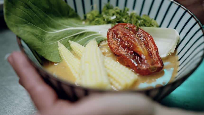 A person holds a striped bowl of hot soup topped with bright green lettuce slices of yellow baby corn and a large roasted red tomato as light reflects on the surface of the liquid
