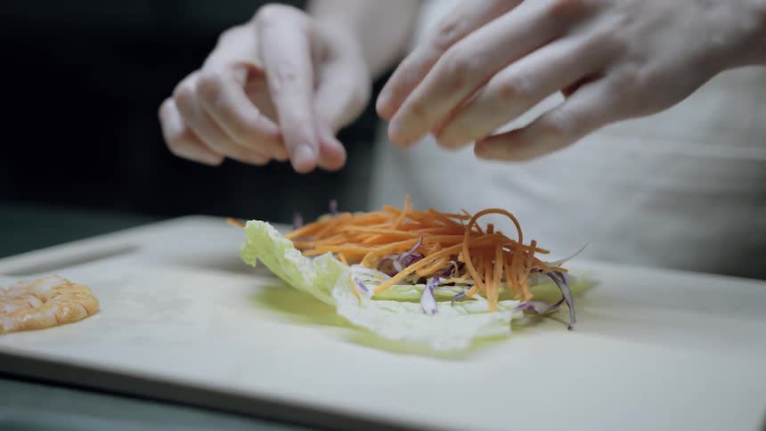 A closeup shot of hands meticulously arranging shredded vegetables into a lettuce leaf on a lightcolored cutting board preparing a fresh food item