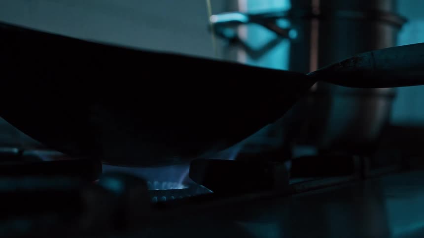 A dark frying pan sits on a stovetop burner with a blue flame as a stream of yellow liquid pours into the pan from above
