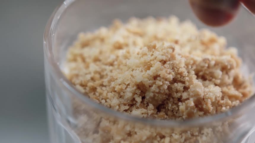 A close view of fingers sprinkling a layer of light brown crumbled granules into a glass