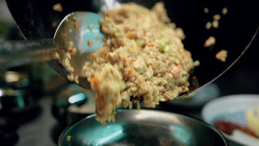 The video shows a closeup of stirfried rice with vegetables and possibly chicken being scooped from a wok with a large spoon the rice is then transferred into a silver bowl
