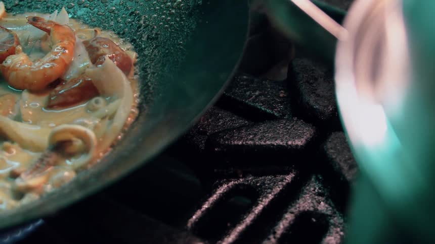 A close view of a dark pan shows shrimp and other seafood cooking in a light colored sauce over a gas burner