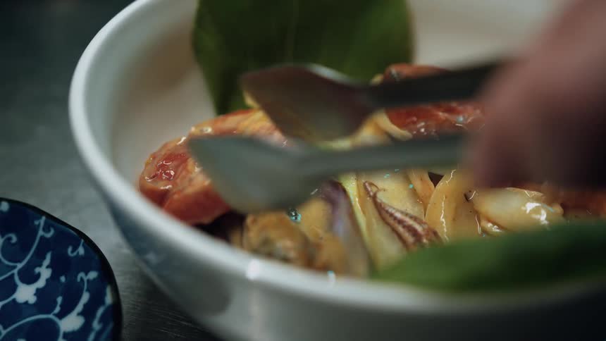 The video shows a closeup of a white bowl filled with a savory dish likely seafood and vegetables being meticulously arranged with tongs the ingredients include mussels sliced tomatoes or sausage and possibly squid suggesting a mixed seafood prepar