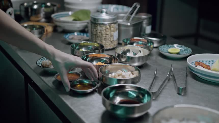 A kitchen prep station displays an array of small metal bowls filled with sauces spices and ingredients as a hand reaches to select one