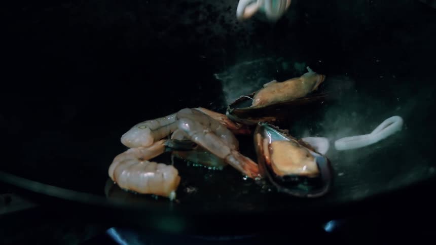 Various pieces of seafood including shrimp mussels and squid rings sizzle in a dark pan over a gas flame as they are cooked against a dark background