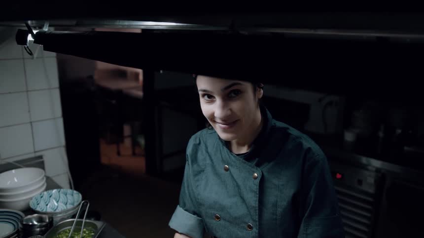 The video shows a person in a dark chefs jacket and a colorful headband standing in what appears to be a professional kitchen they look up towards the camera and smile