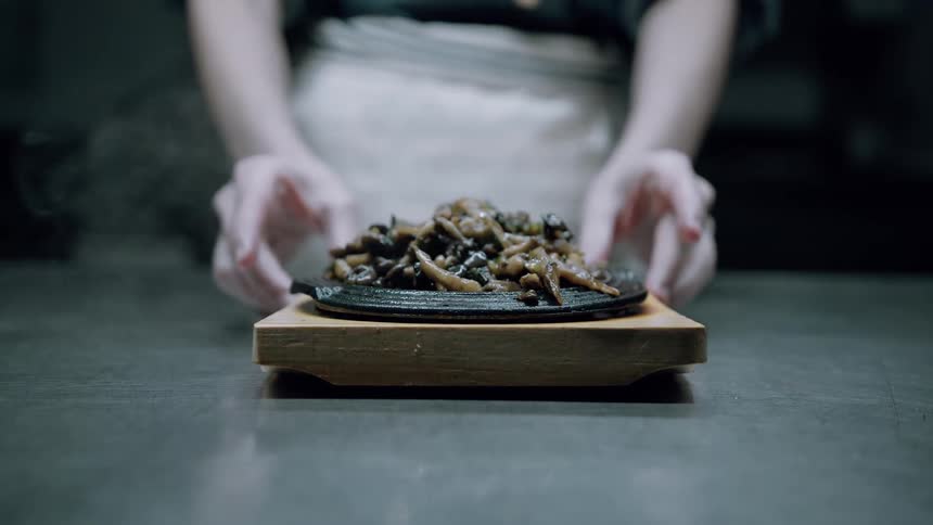 The video shows a closeup of a steaming hot dish likely mushrooms or other small dark vegetables served on a black castiron platter resting on a wooden base a chefs hands wearing an apron are gently placing or presenting the dish on a dark counte