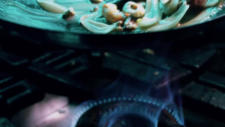 A person moving a sizzling pan of seafood cooks over an open flame releasing steam