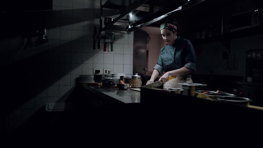 A young chef wearing a dark green chefs coat and a patterned headscarf stands in a dimly lit kitchen chopping ingredients on a metal counter while looking at the viewer and smiling