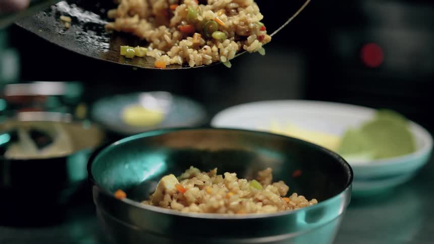 The video shows a closeup of a chef using a large spoon to serve a generous portion of stirfried rice containing vegetables from a wok into a silver bowl the background is blurred focusing attention on the freshly cooked food