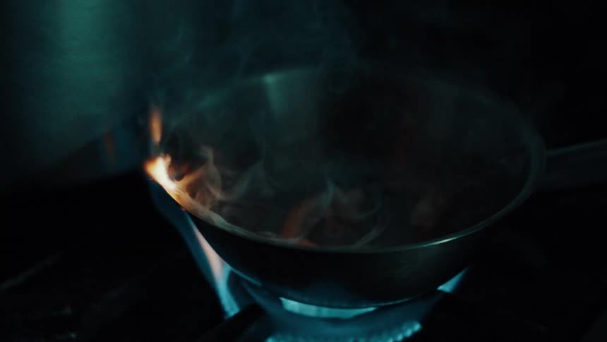 A close view shows a metal pan with a dark exterior and light interior resting on a gas stovetop with a blue flame as ingredients are tossed and briefly ignite with orange flames