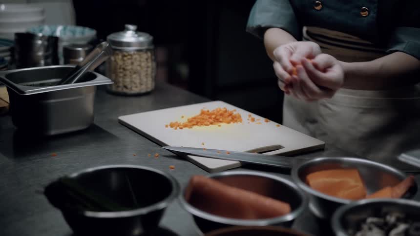 A chef prepares ingredients at a kitchen station with finely chopped vegetables on a cutting board and bowls of ingredients arranged around