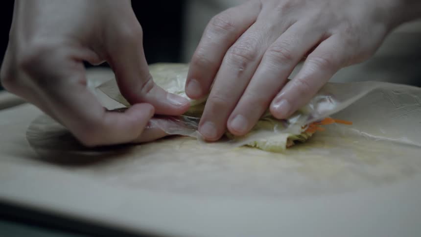 Hands carefully roll vegetables into a rice paper wrapper on a wooden board