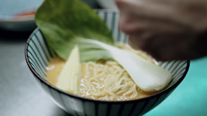A chefs hands add ingredients to a bowl of noodles in a light colored broth placing a large green leaf and yellow corn into the striped bowl