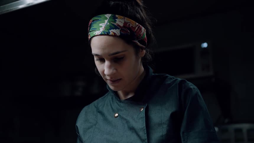 The video shows a closeup of a female chef viewed from the chest up in a dimly lit kitchen she is wearing a dark chefs jacket and a colorful patterned headband and is looking downwards appearing focused on a task
