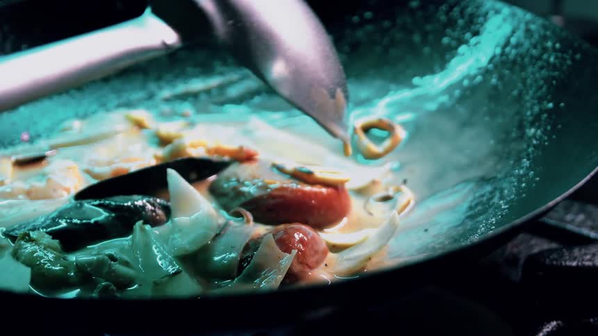 A close view inside a dark pan shows various pieces of seafood including shrimp mussels and squid cooking in a light colored sauce over a blue flame as a large metal spoon stirs the ingredients