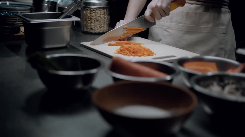 A person in an apron finely chops orange vegetables on a cutting board surrounded by various bowls and cooking tools