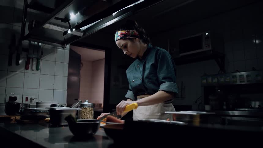 A focused chef in a dark uniform chops food with a knife under warm lighting