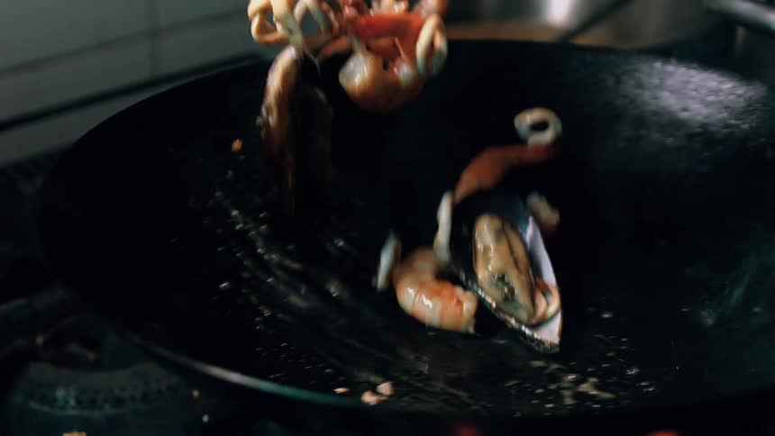 The video shows a closeup of a chef stirfrying seafood in a wok over a gas flame the chef uses tongs to toss mussels shrimp and squid creating motion blur as the ingredients are lifted and fall back into the pan