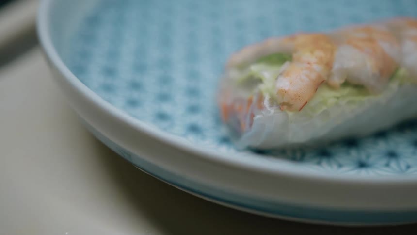 The video shows a closeup of a spring roll being placed into a blue and white patterned bowl a small dark bowl of dipping sauce is then placed next to the spring roll