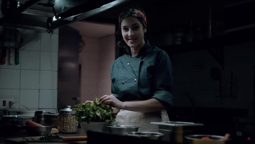 The video shows a cheerful woman in a dark chefs jacket and a colorful headband working in a dimly lit kitchen she is busy preparing food specifically picking through some fresh herbs in a white bowl and she looks up at the camera with a bright smile