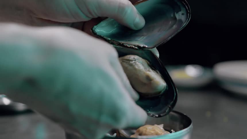 A chef wearing light colored gloves uses their hands to place a steamed mussel from its shell into a small metal bowl of prepared food