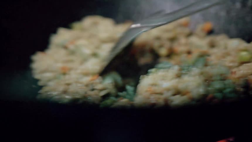 A close view shows fried rice with various vegetables being tossed in a dark pan using a metal spatula with steam rising from the hot food
