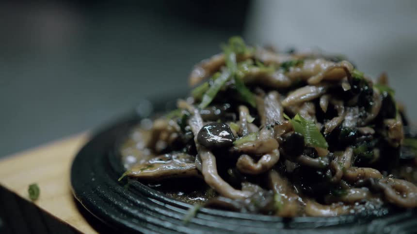 The video displays a closeup of a dark glistening dish likely mushrooms or another vegetable garnished with green herbs served on a round black plate steam or a shimmering effect suggests it is hot and freshly prepared