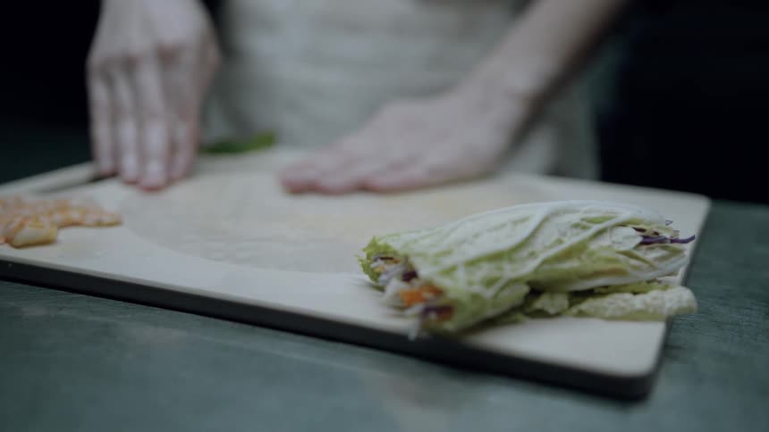 A pair of hands rolls ingredients into a large green lettuce leaf on a wooden cutting board preparing what appears to be a fresh spring roll